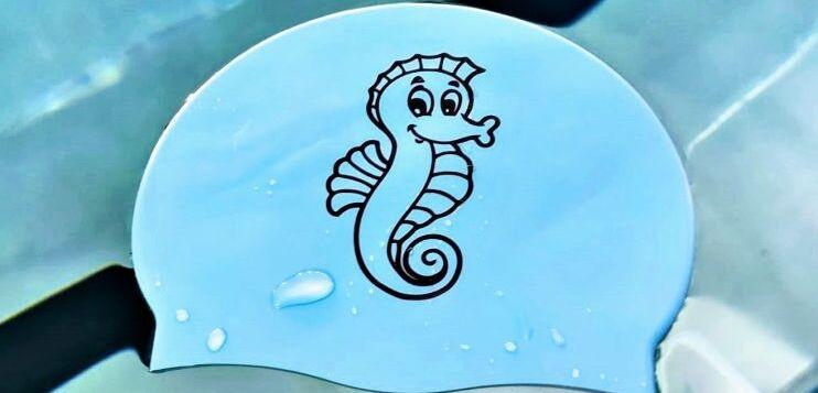 Blauer Schwimmkappen mit einem Seepferdchen-Motiv, auf einer glatten Oberfl&auml;che im Wasser liegend.