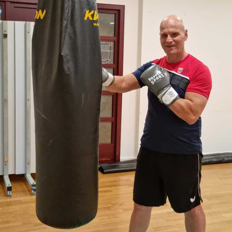 Mann in Boxhandschuhen schl&auml;gt einen schwarzen Boxsack in einem Sportraum mit Holzfu&szlig;boden.