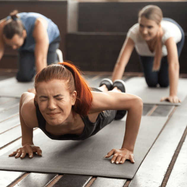 Drei Frauen machen Liegest&uuml;tze auf Fitnessmatten in einem hellen Raum mit Holzfu&szlig;boden. Eine zeigt Anstrengung.