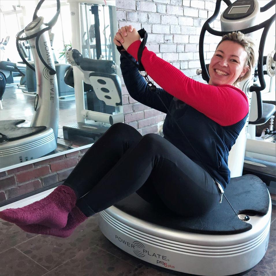 Frau mit l&auml;chelndem Gesicht sitzt auf einer Power Plate und macht eine &Uuml;bung, im Hintergrund Fitnessger&auml;te zu sehen.