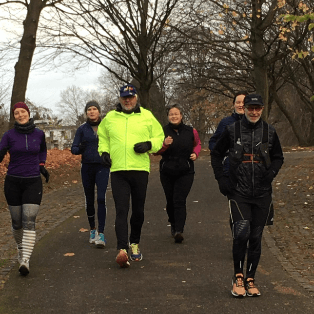 Gruppe von f&uuml;nf Personen joggt auf einem Weg durch einen Park mit herbstlichen B&auml;umen im Hintergrund.