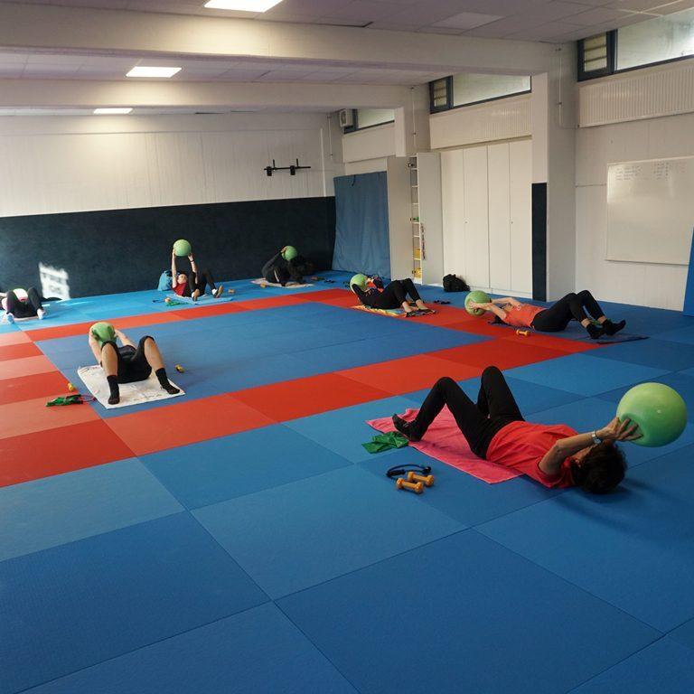 Personen liegen auf Matten und trainieren mit gr&uuml;nen Gymnastikb&auml;llen auf einem blauen Fu&szlig;ballboden.