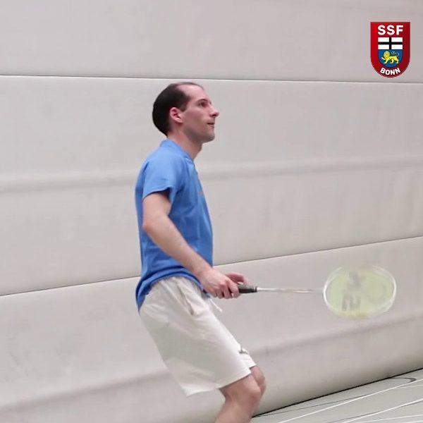 Mann in blauer T-Shirt spielt Badminton in einer Halle, h&auml;lt einen Schl&auml;ger in der Hand und schaut nach oben.