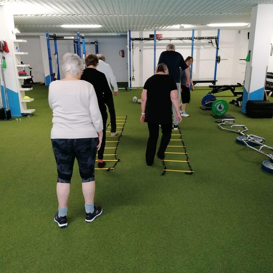 Gruppe von Menschen beim Training mit einer Koordinationsleiter auf einem gr&uuml;nen Sportboden im Fitnessraum.