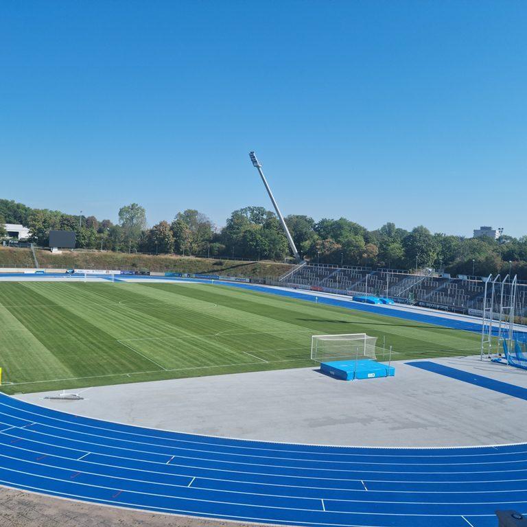 Blick auf ein Sportstadion mit einem gr&uuml;nen Fu&szlig;ballfeld und blauen Laufbahnen unter klarem Himmel.