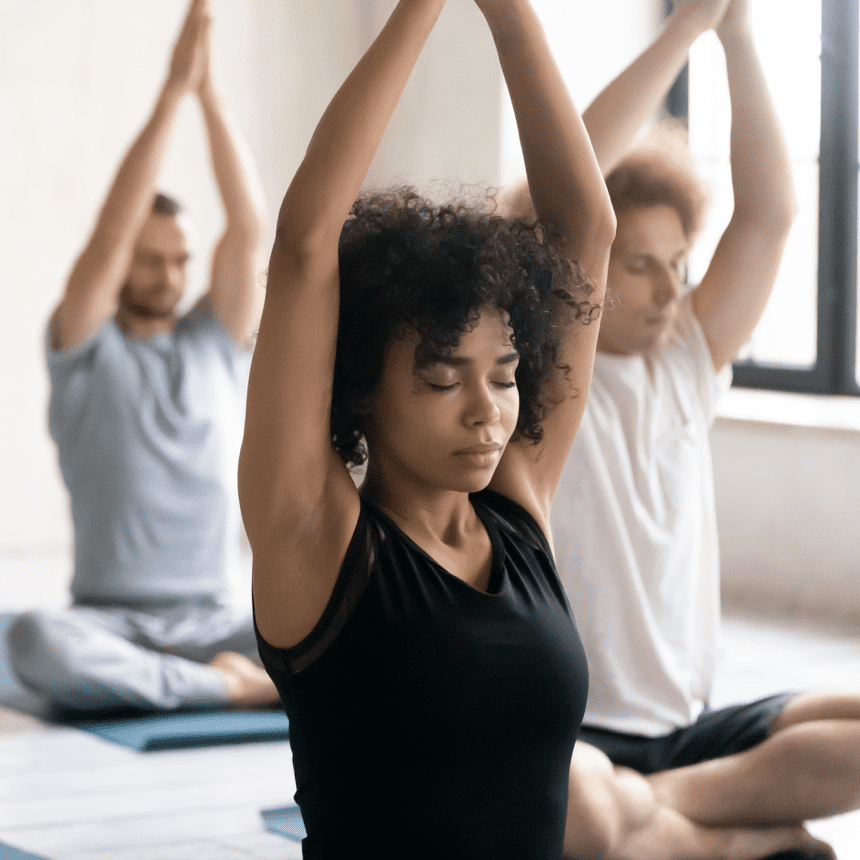 Drei Menschen praktizieren Yoga in einem hellen Raum; eine Frau mit lockigem Haar h&auml;lt die H&auml;nde &uuml;ber dem Kopf.