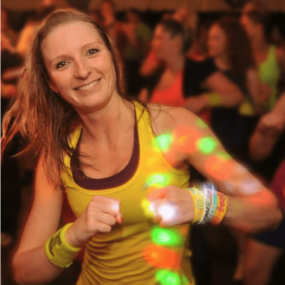 L&auml;chelnde Frau in gelbem Shirt tanzt mit bunten Armb&auml;ndern vor einer Menschenmenge in einer festlichen Atmosph&auml;re.