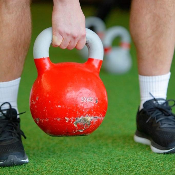 Person hebt eine rote Kettlebell (32 kg) &uuml;ber gr&uuml;nem Bodenbelag in einem Fitnessstudio.