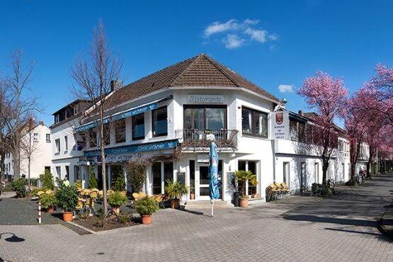 Eckhaus mit Restaurantbeschilderung, umgeben von bl&uuml;henden B&auml;umen und gepflastertem Gehweg, unter blauem Himmel.