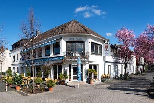 Eckhaus mit Restaurantbeschilderung, umgeben von bl&uuml;henden B&auml;umen und gepflastertem Gehweg, unter blauem Himmel.