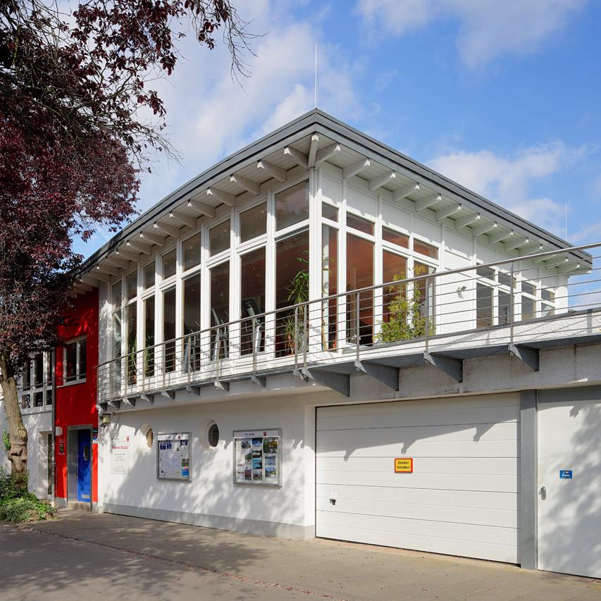 Moderner Bau mit gro&szlig;en Fenstern, roten und wei&szlig;en Fassaden, umgeben von B&auml;umen und einem Gehweg.