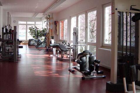 Leerer Fitnessraum mit verschiedenen Ger&auml;ten, gro&szlig;en Fenstern und Blick auf bl&uuml;hende B&auml;ume im Freien.