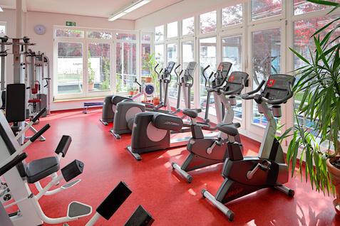 Fitnessraum mit modernen Trainingsger&auml;ten, einschlie&szlig;lich Ergometern, Kraftger&auml;ten und gro&szlig;en Fensterfronten.