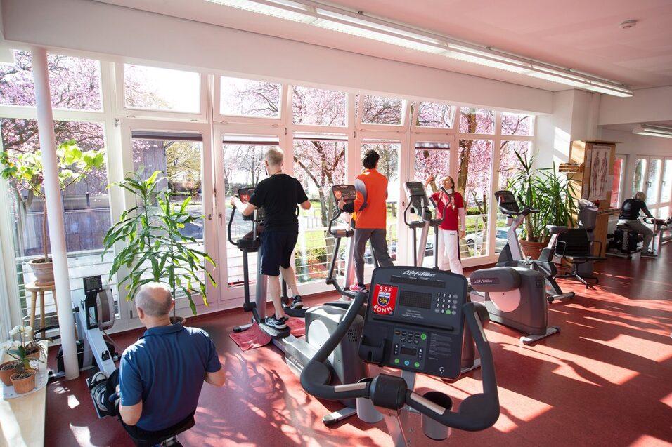 Fitnessraum mit Ger&auml;ten, vier Personen trainieren bei Tageslicht vor gro&szlig;en Fenstern mit bl&uuml;henden B&auml;umen im Hintergrund.