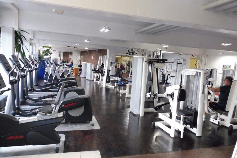 Fitnessstudio mit mehreren Trainingsgeräten und Ausdauermaschinen, während Nutzer aktiv trainieren. Helle, geräumige Umgebung.