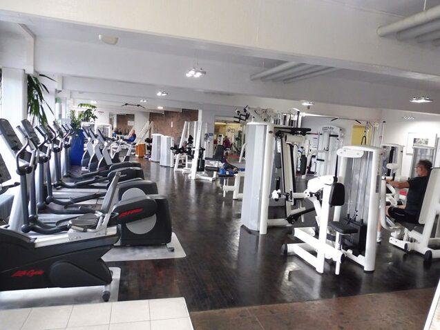 Fitnessstudio mit mehreren Trainingsger&auml;ten und Ausdauermaschinen, w&auml;hrend Nutzer aktiv trainieren. Helle, ger&auml;umige Umgebung.