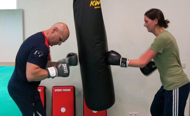 Zwei Personen trainieren Boxen, eine schl&auml;gt gegen einen schwarzen Boxsack, die andere beobachtet.