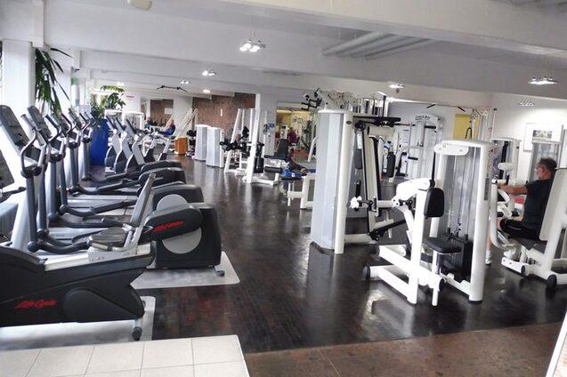 Fitnessstudio mit mehreren Trainingsger&auml;ten und Ausdauermaschinen, w&auml;hrend Nutzer aktiv trainieren. Helle, ger&auml;umige Umgebung.