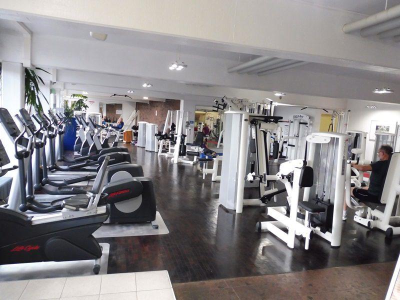 Fitnessstudio mit mehreren Trainingsgeräten und Ausdauermaschinen, während Nutzer aktiv trainieren. Helle, geräumige Umgebung.