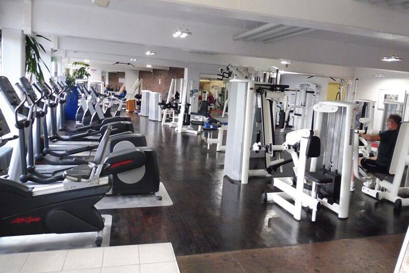 Fitnessstudio mit mehreren Trainingsger&auml;ten und Ausdauermaschinen, w&auml;hrend Nutzer aktiv trainieren. Helle, ger&auml;umige Umgebung.