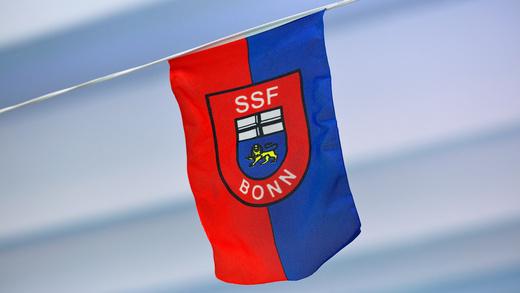 Flagge des SSF Bonn mit rotem und blauem Design, Wappen und Schriftzug "SSF BONN" in der Mitte.