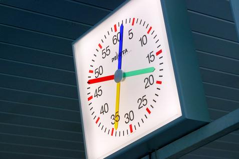 Analog-Uhr mit bunten Zeigern, die auf 3:35 Uhr zeigt, an einer Wand montiert.