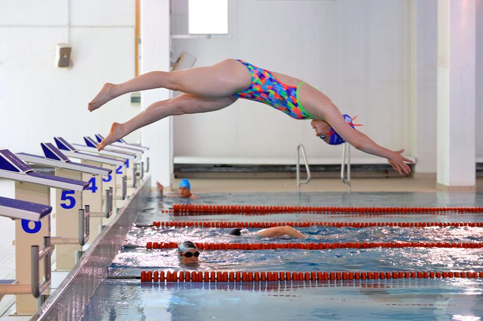 Schwimmerin springt in einem Sportbecken von der Startbl&ouml;cken, w&auml;hrend ein weiterer Schwimmer im Wasser schwimmt.
