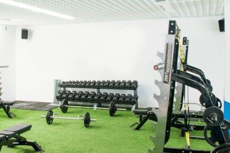 Fitnessraum mit Hanteln, Trainingsb&auml;nken und Kraftger&auml;ten auf gr&uuml;nem Kunstrasenboden vor wei&szlig;en W&auml;nden.