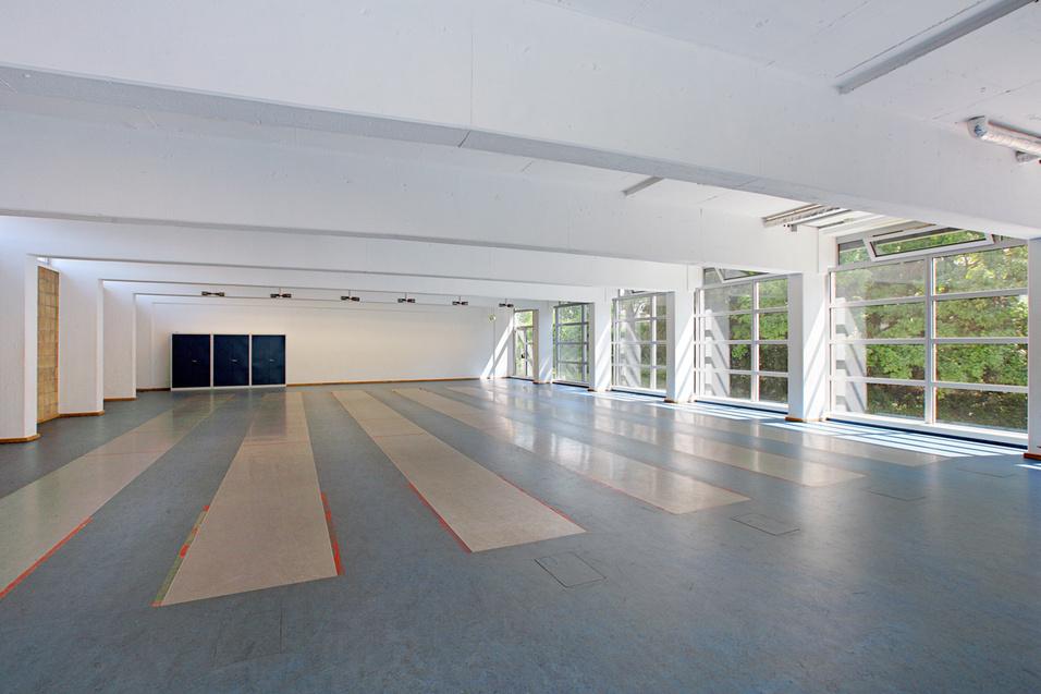 Leere Sporthalle mit gro&szlig;en Fenstern, grauem Boden und mehreren Bahnen f&uuml;r Wettk&auml;mpfe oder Trainings.