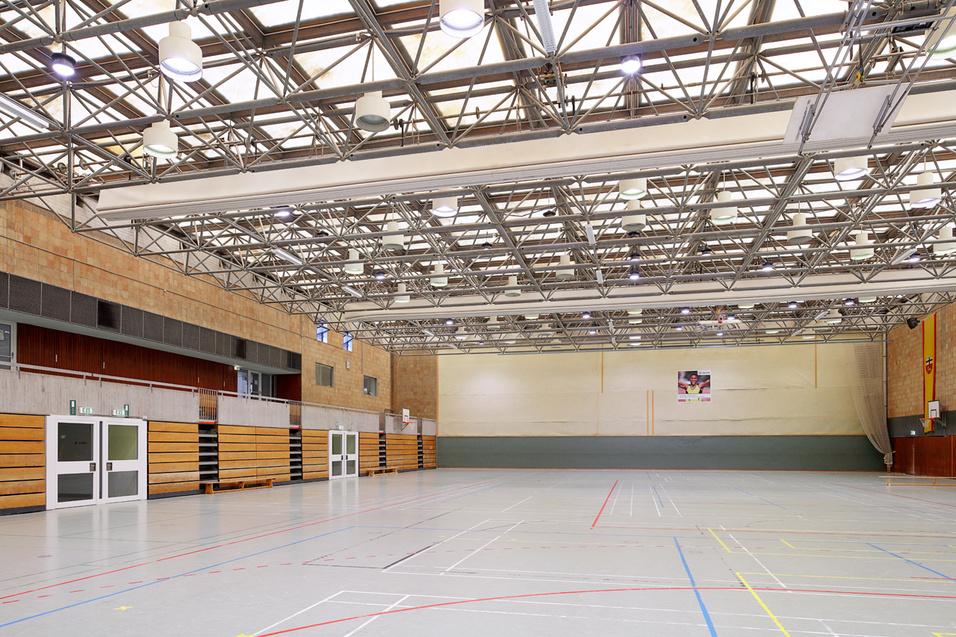 Helle Sporthalle mit Holzw&auml;nden, gro&szlig;en Fenstern und einem leeren, markierten Sportboden. Deckenleuchten sind sichtbar.