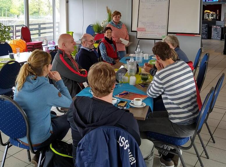 Gruppendiskussion in einem Caf&eacute; mit Personen um einen Tisch, einer Frau, die eine Pr&auml;sentation gibt, und Getr&auml;nken auf dem Tisch.