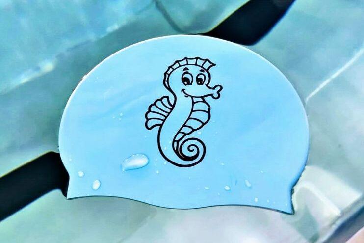 Schwimmkappe in Blau mit einem schwarzen Seepferdchen-Motiv auf einer glitschigen Oberfl&auml;che.