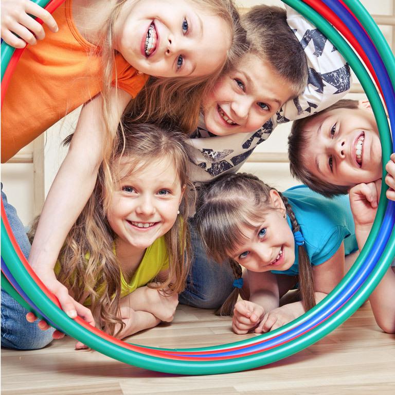 F&uuml;nf Kinder l&auml;cheln fr&ouml;hlich und schauen durch einen bunten Hula-Hoop-Reifen auf einem Holzfu&szlig;boden.