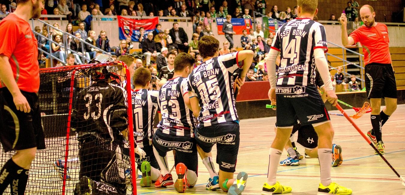 Szenario eines Floorball-Spiels mit Spielern in Schwarz und Wei&szlig; vor Tor und jubelnden Zuschauern im Hintergrund.