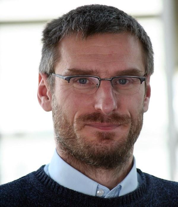 Portr&auml;t eines Mannes mit Brille, kurzer grauer Haartracht und Bart, l&auml;chelt vor einem unscharfen Hintergrund.