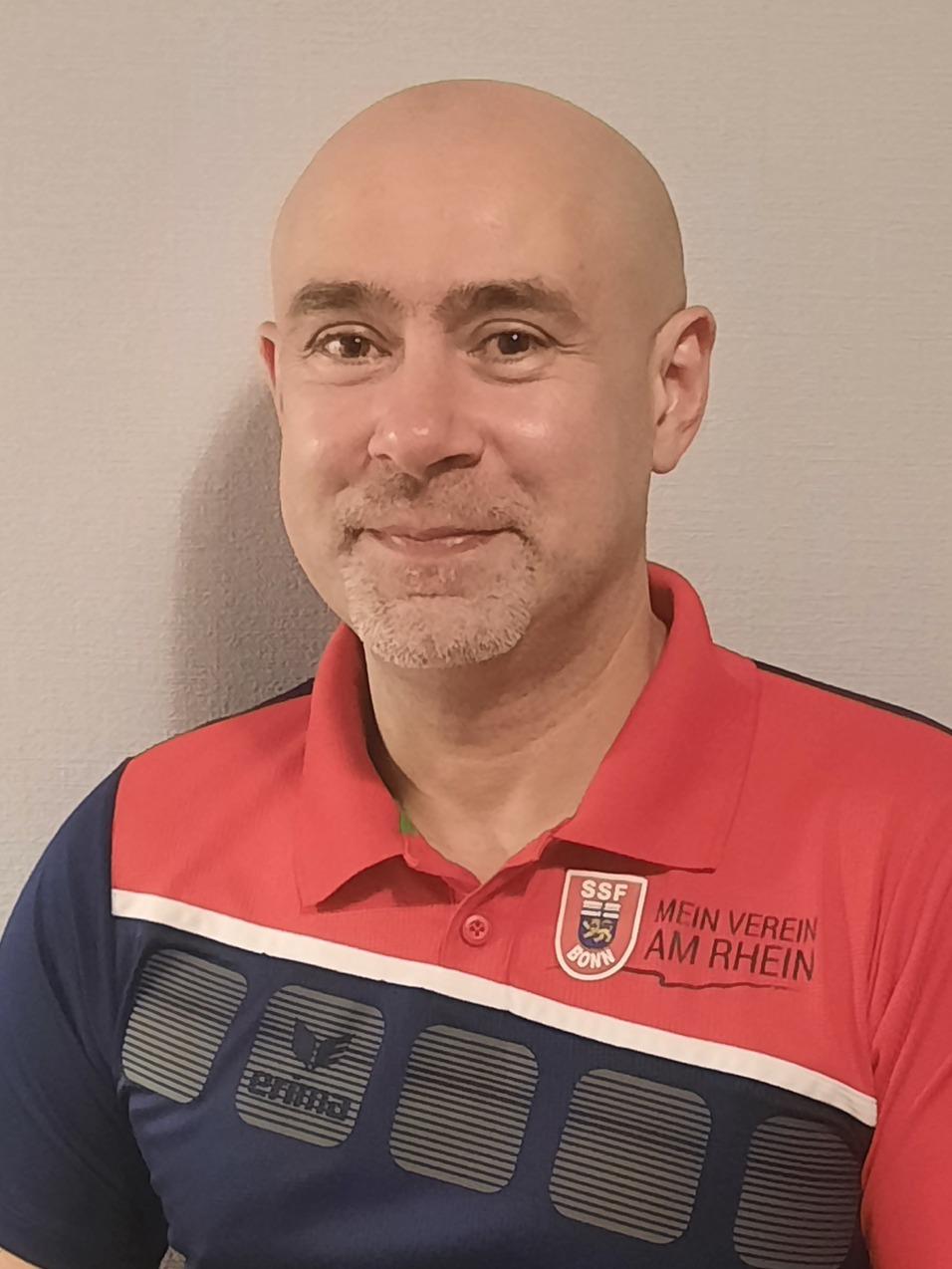 Mann mit Glatze in rotem Polohemd mit blauem Muster steht vor einer hellen Wand.