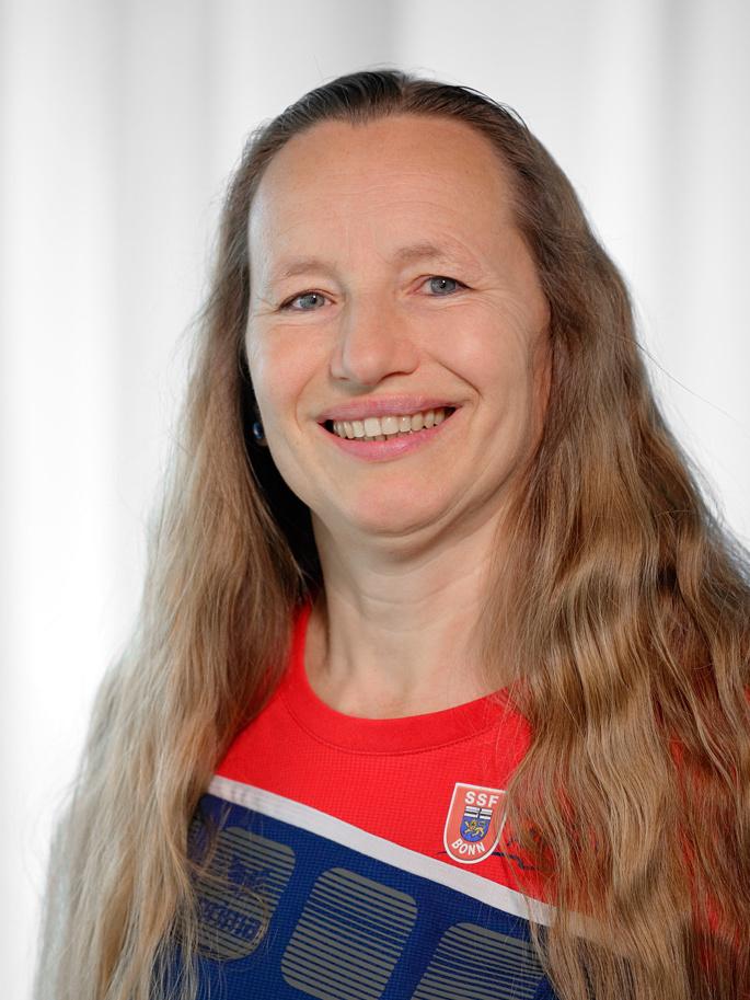Portrait einer l&auml;chelnden Frau mit langen, blonden Haaren, im roten Sporttrikot vor neutralem Hintergrund.