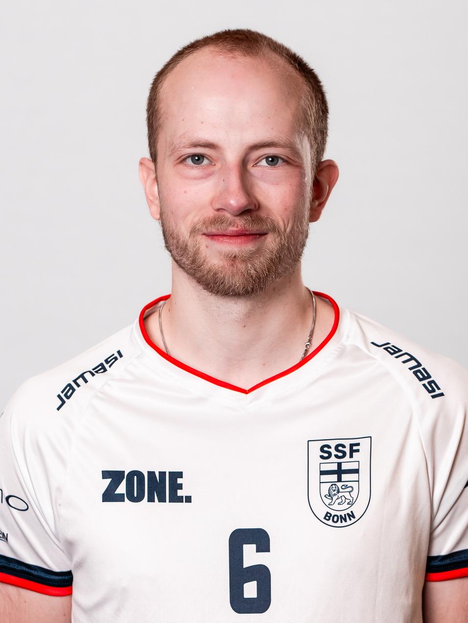 Portr&auml;t eines Mannes in einem wei&szlig;en Trikot mit der Nummer 6, l&auml;chelnd vor einem neutralen Hintergrund.