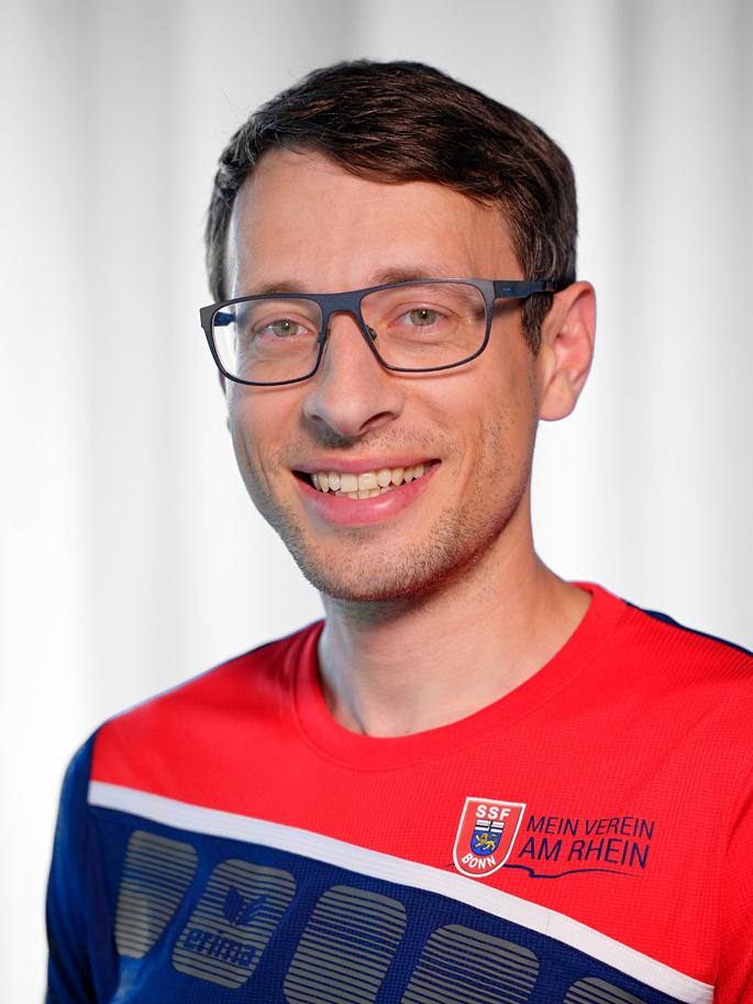 Portr&auml;t eines l&auml;chelnden Mannes mit Brille, der ein rotes Sporttrikot mit blau-wei&szlig;en Details tr&auml;gt.