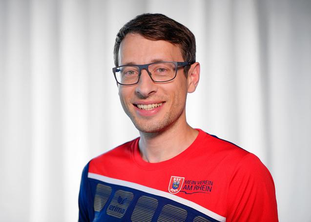 Portr&auml;t eines l&auml;chelnden Mannes mit Brille, der ein rotes Sporttrikot mit blau-wei&szlig;en Details tr&auml;gt.