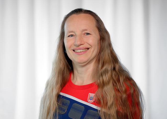 Portrait einer l&auml;chelnden Frau mit langen, blonden Haaren, im roten Sporttrikot vor neutralem Hintergrund.