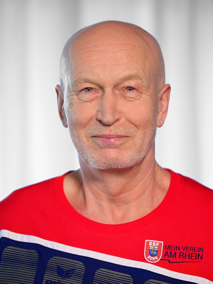 Portrait eines l&auml;chelnden Mannes in einem rot-blauen Sportshirt mit Vereinsaufschrift, vor hellem Hintergrund.