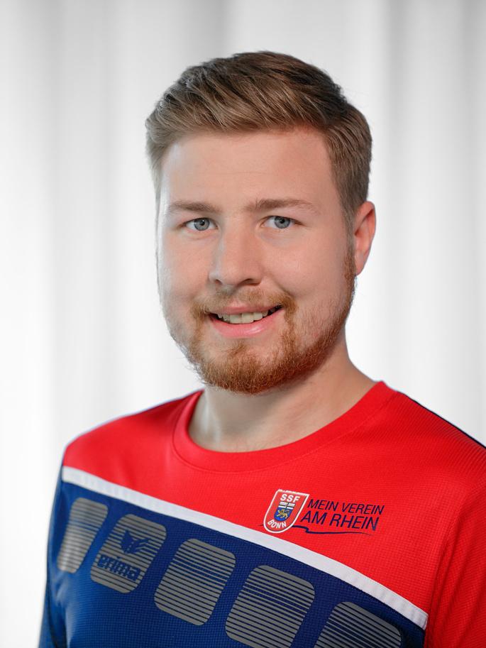 Portr&auml;t eines l&auml;chelnden Mannes mit blonden Haaren und Bart, tr&auml;gt ein rotes Sporttrikot mit blauem Design.