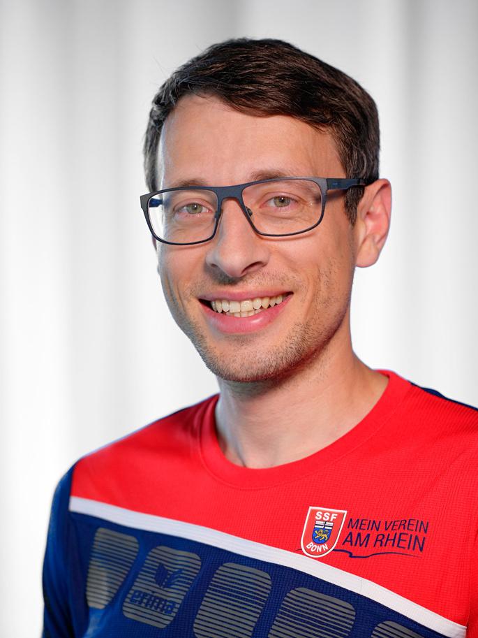 Portr&auml;t eines l&auml;chelnden Mannes mit Brille, der ein rotes Sporttrikot mit blau-wei&szlig;en Details tr&auml;gt.