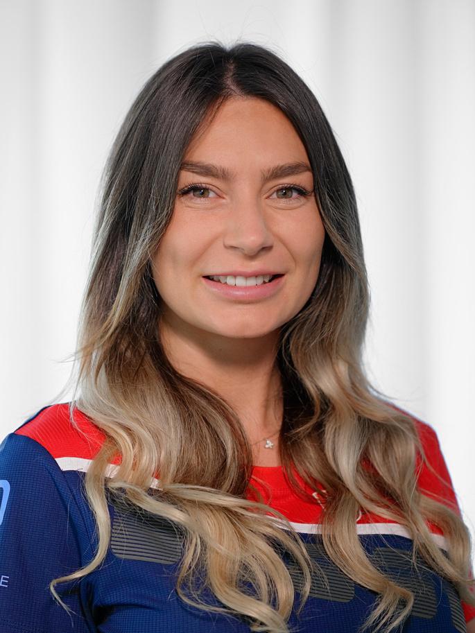 Portrait einer l&auml;chelnden Frau mit langen, gewellten Haaren, im sportlichen Outfit vor einem hellen Hintergrund.