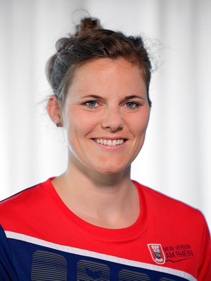 Portr&auml;t einer l&auml;chelnden Frau mit einem Dutt, die ein rotes Sporttrikot tr&auml;gt und vor einem hellen Hintergrund steht.