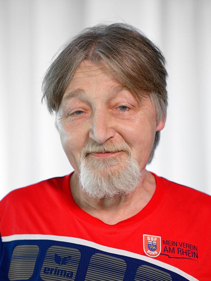 Portr&auml;t eines &auml;lteren Mannes mit Bart, l&auml;chelnd, in rotem Sporttrikot mit Vereinslogo auf wei&szlig;em Hintergrund.