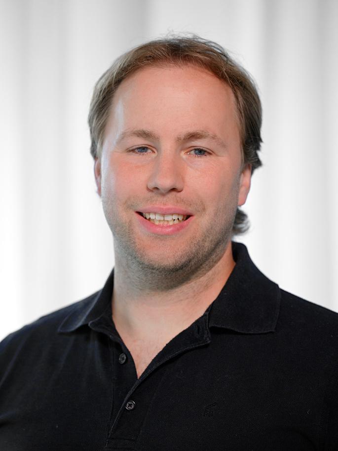 Portr&auml;t eines l&auml;chelnden Mannes mit kurzem, blonden Haar, tr&auml;gt ein schwarzes Polo-Shirt vor einem hellen Hintergrund.