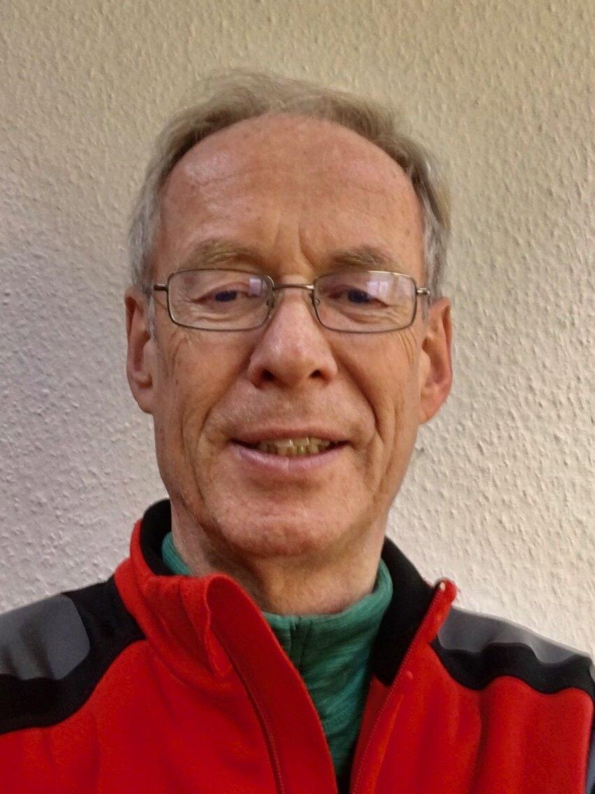 &Auml;lterer Mann mit Brille und grauen Haaren tr&auml;gt einen roten und schwarzen Pullover vor einer hellen Wand.