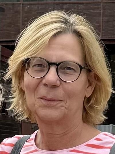 Nahaufnahme einer Frau mit blonden, lockigen Haaren und einer Brille, die leicht l&auml;chelt. Sie tr&auml;gt ein gestreiftes Oberteil.